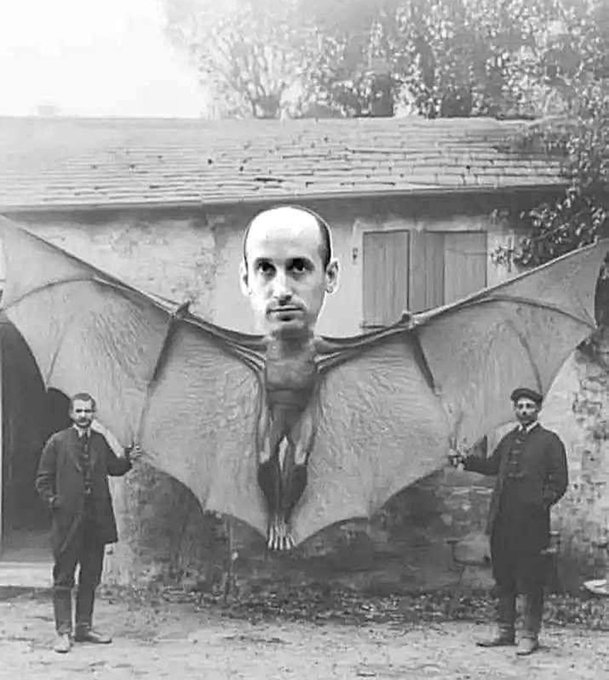 Black-and-white photograph depicts two men in early 20th-century attire standing on a cobblestone path in front of a stone building with wooden shutters. They hold outstretched wings of a large bat-like creature positioned centrally. The creatures body is furry with clawed feet and the head is that of a bald middle-aged man with a neutral expression seamlessly edited onto it forming a surreal hybrid figure.