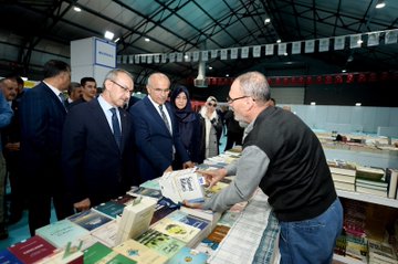 First image shows a large group of formally dressed men and women in suits and dresses standing together cutting a red ribbon in front of a banner reading Malatya Anadolu Kitap Fuarı with Turkish flags and exhibition hall setting. Second image depicts a crowd of attendees including men in suits and women in hijabs applauding around a young girl in red and white traditional Anatolian costume with arms raised on a tiled floor near exhibition banners. Third image features four men in suits and a woman in hijab examining and holding books on a table filled with stacked books in an exhibition booth under hanging flags. Fourth image captures a red-haired elderly woman in a red outfit signing books at a table surrounded by stacks of books while a young man in green shirt leans in attentively with other attendees and exhibition displays in the background.