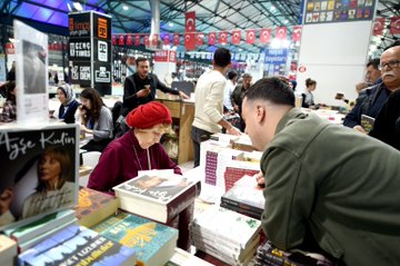 First image shows a large group of formally dressed men and women in suits and dresses standing together cutting a red ribbon in front of a banner reading Malatya Anadolu Kitap Fuarı with Turkish flags and exhibition hall setting. Second image depicts a crowd of attendees including men in suits and women in hijabs applauding around a young girl in red and white traditional Anatolian costume with arms raised on a tiled floor near exhibition banners. Third image features four men in suits and a woman in hijab examining and holding books on a table filled with stacked books in an exhibition booth under hanging flags. Fourth image captures a red-haired elderly woman in a red outfit signing books at a table surrounded by stacks of books while a young man in green shirt leans in attentively with other attendees and exhibition displays in the background.