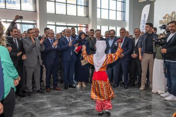 First image shows a large group of formally dressed men and women in suits and dresses standing together cutting a red ribbon in front of a banner reading Malatya Anadolu Kitap Fuarı with Turkish flags and exhibition hall setting. Second image depicts a crowd of attendees including men in suits and women in hijabs applauding around a young girl in red and white traditional Anatolian costume with arms raised on a tiled floor near exhibition banners. Third image features four men in suits and a woman in hijab examining and holding books on a table filled with stacked books in an exhibition booth under hanging flags. Fourth image captures a red-haired elderly woman in a red outfit signing books at a table surrounded by stacks of books while a young man in green shirt leans in attentively with other attendees and exhibition displays in the background.