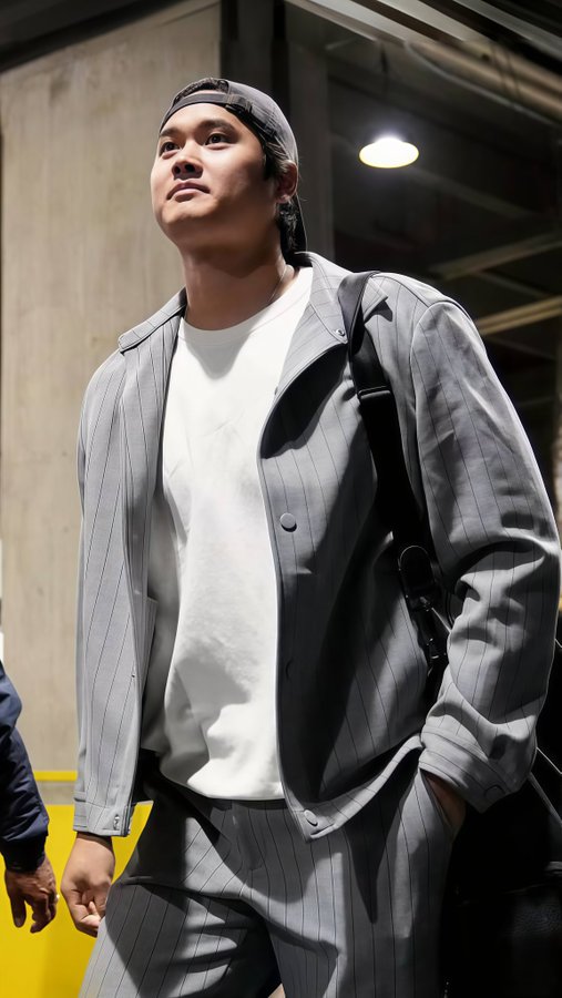 Young Asian man with short black hair wearing a black cap tilted back, white short-sleeve t-shirt, open gray pinstripe button-up shirt, matching gray pants, and black backpack slung over one shoulder, standing with hands in pockets and looking upward slightly in an indoor space with concrete walls, overhead fluorescent lights, and a yellow section visible nearby.