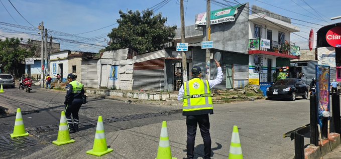 Two police officers in black uniforms and yellow high-visibility vests stand on a paved street directing traffic, one raising a hand while the other stands nearby. Orange traffic cones are placed along the road edges and near a black SUV parked on the side. In the background, there are several low-rise buildings with signs including a green one reading SALON and a traffic light visible. A motorcycle with a rider in a helmet is positioned near the cones, and pedestrians are visible in the distance. The setting appears to be an urban residential area with trees and utility poles.
