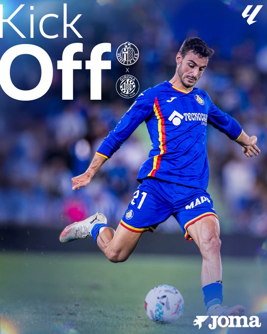 A soccer player in a blue Joma kit with number 21 and sponsor logos including Tecnocasa and MAD stands on a green field kicking a white soccer ball mid-air during kickoff. The background shows a stadium with blurred stands and field markings. Overlaid text reads Kick Off in large white letters with Getafe and Girona club crests. The player has short dark hair and is in dynamic motion with one leg extended.