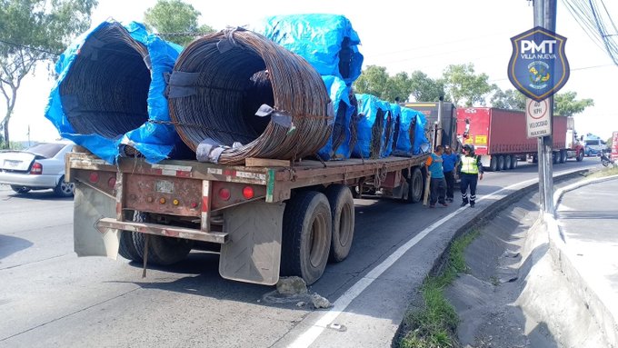 Red semi-trailer truck loaded with multiple large blue plastic-wrapped coils of steel rebar, parked on the edge of a roadside with a slight uphill slope, two workers in yellow safety vests and helmets standing nearby, a white car in the background, a blue sign reading P.M.T. VILLA NUEVA with emblem on a pole, and another truck visible further along the road under clear daytime sky.