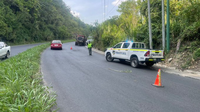 Roadside scene on a curved highway with green hills and vegetation in background shows a white Provial truck with yellow markings and flashing lights parked behind a large red tanker truck partially off the road obstructing the right lane a person in high-visibility vest stands near the tanker orange traffic cones mark the area a white car and green truck are visible nearby a yellow rope or barrier extends from the scene.