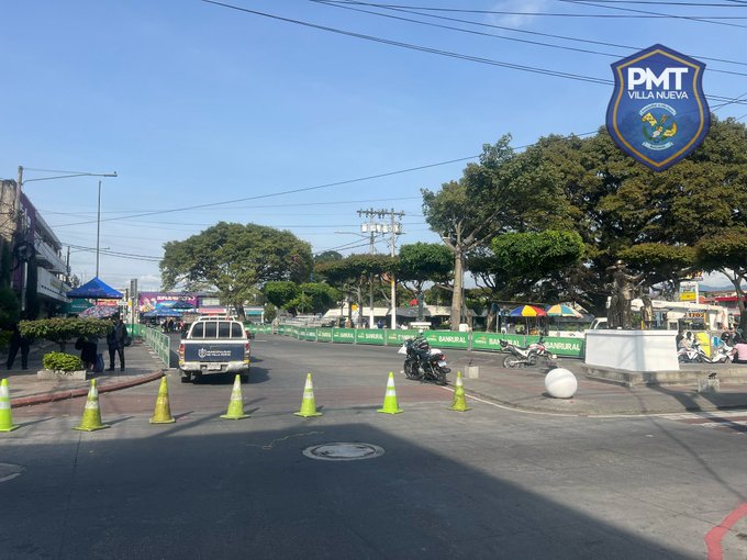 Street scene with yellow traffic cones arranged to block access, police van parked nearby with PMT Villa Nueva emblem visible on it and a large sign, motorcycles and other vehicles present, trees and urban buildings in background under clear sky.