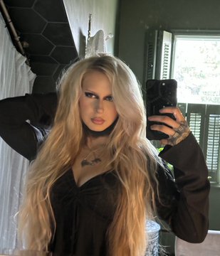 A woman with long wavy blonde hair and bold eye makeup stands in a green-tiled bathroom with wooden shutters and plants in the background. She wears a black low-cut dress with a necklace and holds a black iPhone in her tattooed hand for a mirror selfie. The two similar images show her posing with hands raised near her face emphasizing her outfit and expression.