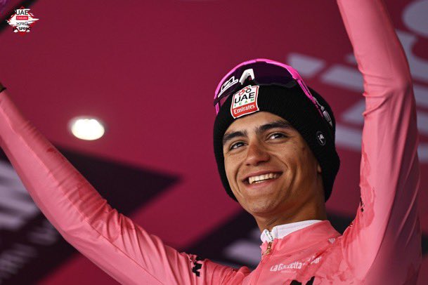A young male cyclist with short dark hair wearing a black UAE Team Emirates beanie hat smiles broadly while raising both arms in celebration on a dimly lit indoor stage with red and pink lighting. He is dressed in a pink long-sleeve cycling jersey with white accents and sponsor logos including UAE and Lidl visible on the chest and sleeves. The background shows a blurred crowd and event branding.
