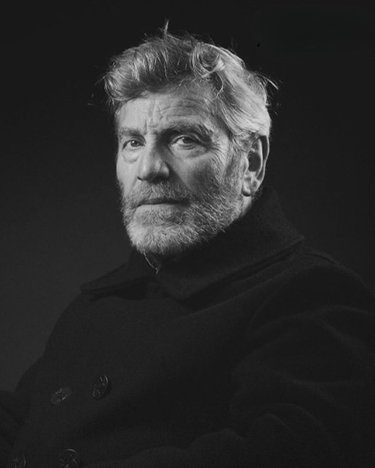 Black-and-white portrait of Tcheky Karyo, a man with curly gray hair and beard, wearing a dark coat with buttons, facing forward against a dark background.