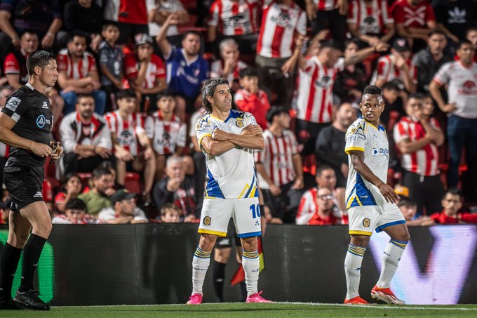 Two soccer players in white and blue striped uniforms with red accents stand on the green field, one with arms crossed wearing number 10 jersey, the other nearby, a referee in black uniform positioned beside them, stadium crowd in background wearing red and white supporter attire filling the stands.