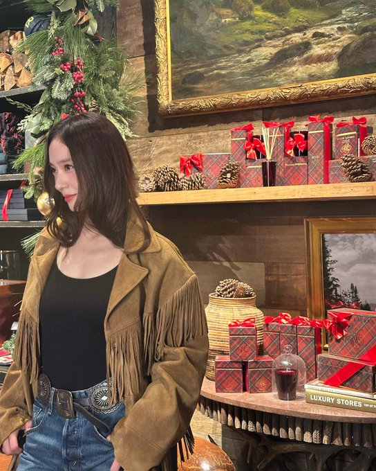 First image shows a woman with long dark hair wearing a fringed tan leather jacket over a black tank top paired with blue jeans a wide belt and brown boots standing in a rustic wooden store interior with saddles fishing nets antlers and a stable sign on the wall behind her holding a brown crossbody bag. Second image features the same woman in a yellow fringed jacket blue jeans and black boots posing near a large decorated Christmas tree with golden ornaments string lights and a wooden box labeled Ralph Lauren in a festive outdoor-like setting. Third image depicts her in a brown fringed leather jacket black tank top blue jeans and boots near holiday decorations including a Christmas tree garlands red gift boxes pinecones and paintings in a luxury store environment with a round table and glass dome.