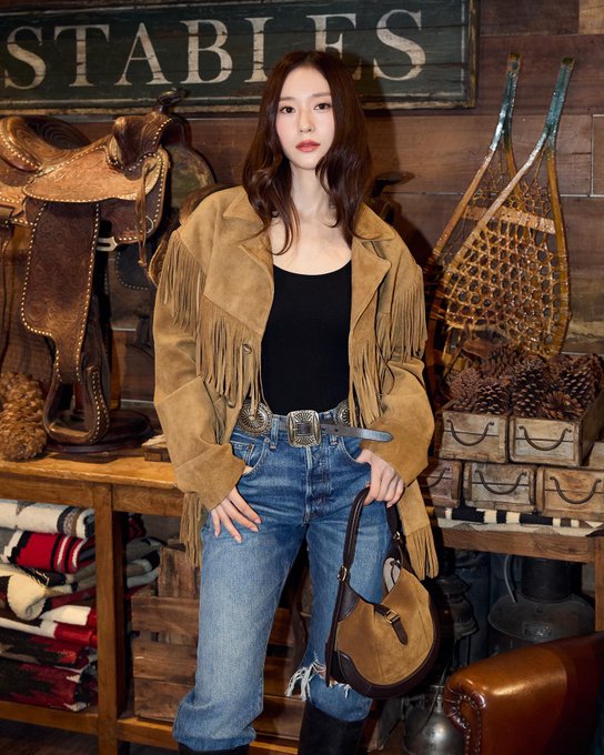 First image shows a woman with long dark hair wearing a fringed tan leather jacket over a black tank top paired with blue jeans a wide belt and brown boots standing in a rustic wooden store interior with saddles fishing nets antlers and a stable sign on the wall behind her holding a brown crossbody bag. Second image features the same woman in a yellow fringed jacket blue jeans and black boots posing near a large decorated Christmas tree with golden ornaments string lights and a wooden box labeled Ralph Lauren in a festive outdoor-like setting. Third image depicts her in a brown fringed leather jacket black tank top blue jeans and boots near holiday decorations including a Christmas tree garlands red gift boxes pinecones and paintings in a luxury store environment with a round table and glass dome.