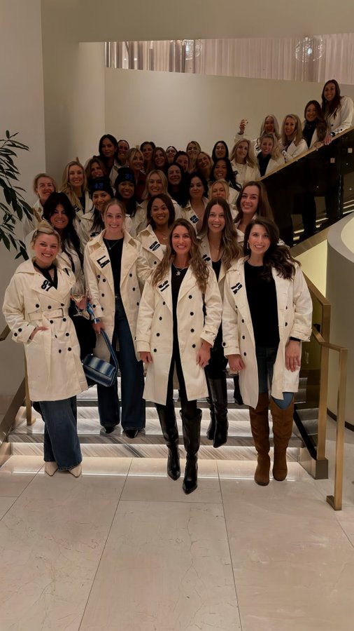 Numerous women stand and sit on wide stairs in a brightly lit modern lobby with glass railings and neutral tones. They wear identical beige trench coats with black belts, some open and some tied, over various outfits including jeans and dresses. Footwear includes knee-high boots in black and brown. Handbags in blue and black are held by some. Expressions are smiling and neutral, with diverse hairstyles and skin tones. The group arrangement is casual and clustered.