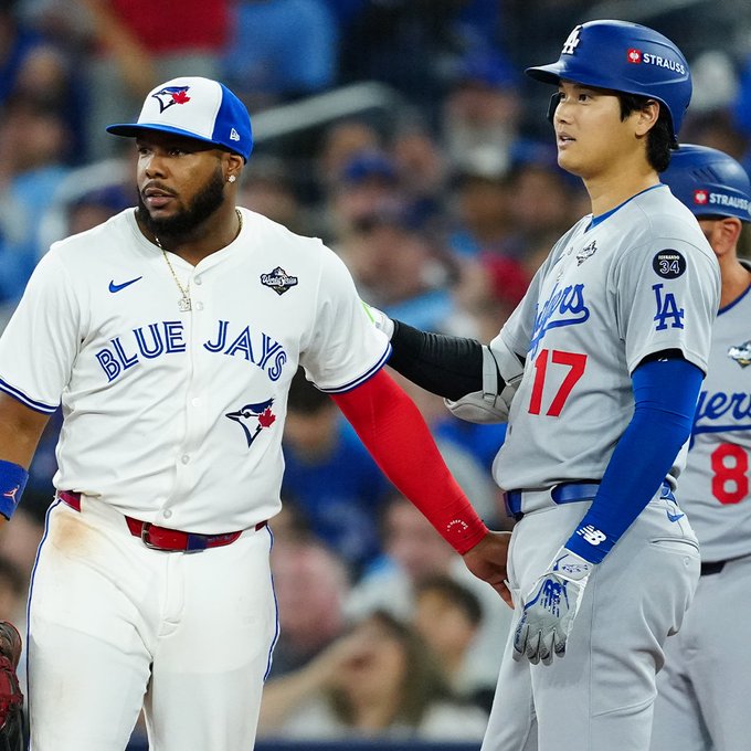 Vladimir Guerrero Jr. and Shohei Ohtani interact at first base during World Series Game 2. Guerrero Jr. is wearing a white Toronto Blue Jays jersey, and Ohtani is wearing a gray Los Angeles Dodgers jersey.