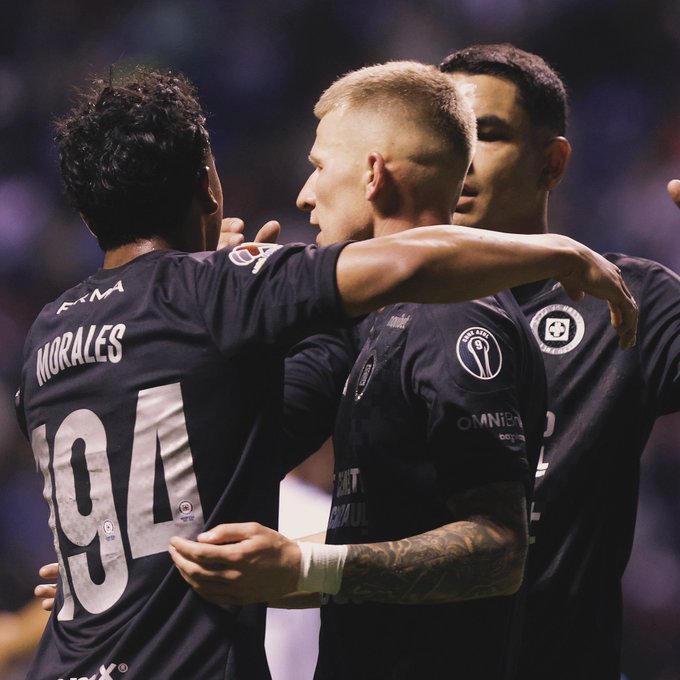 Three male soccer players in dark blue Cruz Azul jerseys with number 104 and name Morales on one back celebrate by hugging on a stadium field during a match, one with blond hair facing forward, another with dark hair turned sideways, and a third with light skin and tattoos on arm visible, stadium lights and crowd in blurred background.