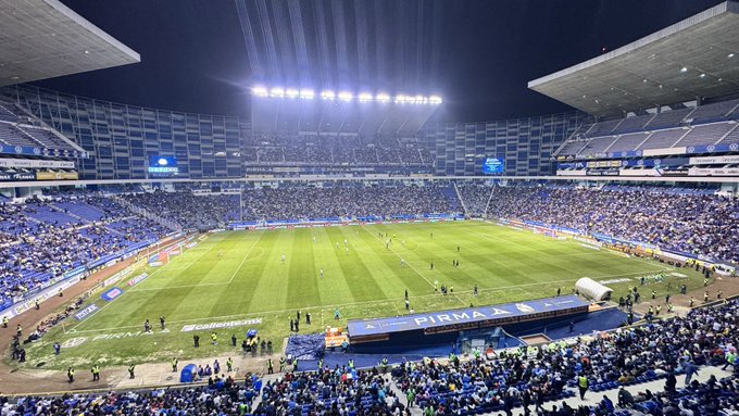 Aerial view of a large modern soccer stadium at night under bright floodlights, filled with a crowd of spectators mostly wearing blue clothing in the stands surrounding the green field, with team benches and a scoreboard visible on the field, and the word PINMA displayed on a banner.