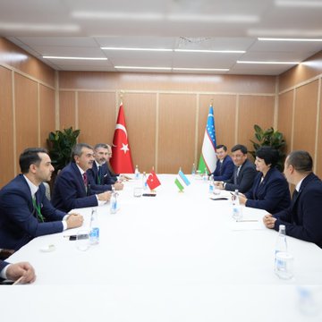 First image shows a formal conference room with wooden paneling and a long table covered in white cloth, where several suited officials sit engaged in discussion, Turkish and Uzbek flags positioned at the ends, documents and notebooks on the table, a graphic overlay announcing the protocol signing in Turkish with ministry logos. Second image depicts a similar meeting setup with more officials around the table, water bottles and papers present, flags of Turkey and Uzbekistan flanking the scene, plants in the background for decoration. Third image captures two officials, a man in a dark suit with a red tie holding a red folder and a woman in a blue suit holding a blue folder, shaking hands in front of wooden walls with Turkish and Uzbek flags on stands.