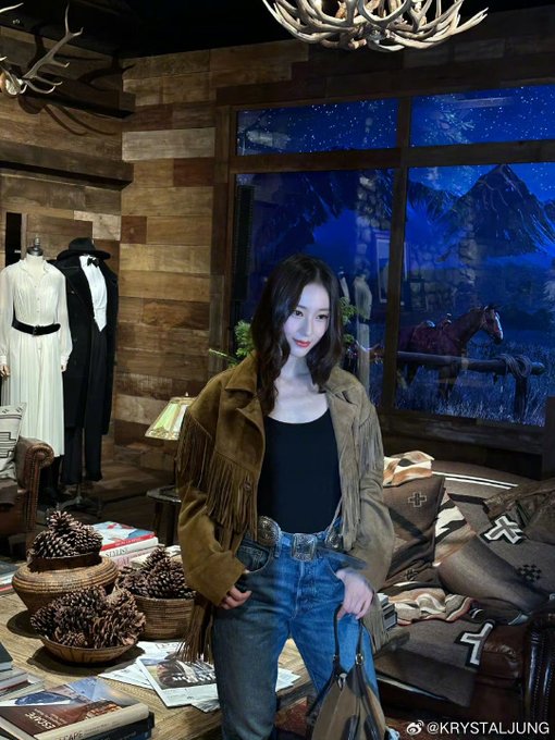Woman with long dark hair wearing fringe suede jacket black top blue jeans and black boots stands near yellow LAURA sign with golden holiday lights and trees in background. Woman in similar outfit poses in wooden stable area with STABLES sign saddles cowboy boots and rustic decor. Woman in fringe jacket black top jeans and boots stands near wooden shelves with saddles antlers and STABLES sign in rustic setting. Woman in fringe suede jacket black top jeans and boots poses in wooden interior with antlers display cases white dress form and window view of blue sky.