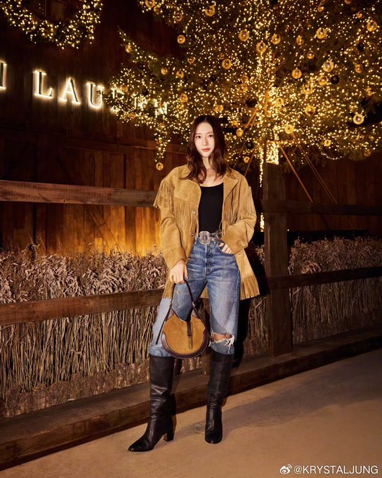 Woman with long dark hair wearing fringe suede jacket black top blue jeans and black boots stands near yellow LAURA sign with golden holiday lights and trees in background. Woman in similar outfit poses in wooden stable area with STABLES sign saddles cowboy boots and rustic decor. Woman in fringe jacket black top jeans and boots stands near wooden shelves with saddles antlers and STABLES sign in rustic setting. Woman in fringe suede jacket black top jeans and boots poses in wooden interior with antlers display cases white dress form and window view of blue sky.
