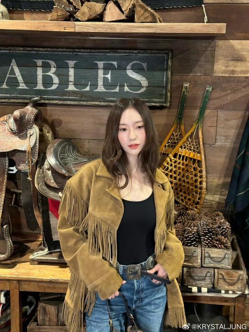 Woman with long dark hair wearing fringe suede jacket black top blue jeans and black boots stands near yellow LAURA sign with golden holiday lights and trees in background. Woman in similar outfit poses in wooden stable area with STABLES sign saddles cowboy boots and rustic decor. Woman in fringe jacket black top jeans and boots stands near wooden shelves with saddles antlers and STABLES sign in rustic setting. Woman in fringe suede jacket black top jeans and boots poses in wooden interior with antlers display cases white dress form and window view of blue sky.