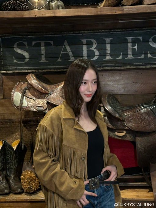 Woman with long dark hair wearing fringe suede jacket black top blue jeans and black boots stands near yellow LAURA sign with golden holiday lights and trees in background. Woman in similar outfit poses in wooden stable area with STABLES sign saddles cowboy boots and rustic decor. Woman in fringe jacket black top jeans and boots stands near wooden shelves with saddles antlers and STABLES sign in rustic setting. Woman in fringe suede jacket black top jeans and boots poses in wooden interior with antlers display cases white dress form and window view of blue sky.