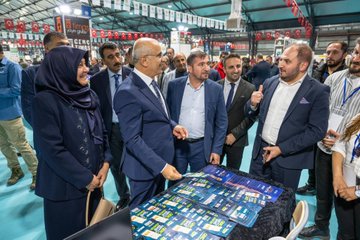 First image displays a group of formally dressed men and women including some in headscarves standing around a table covered with stacks of blue-covered books at an indoor event space with Turkish flags and banners overhead. Second image shows three men in suits one holding a book posing in front of a banner for a book model event with a screen and books on a table. Third image features a group of young men and women some in headscarves standing together at a booth labeled for second category fair with coffee cups and displayed items on the table. Fourth image depicts a line of children in colorful traditional Turkish attire including embroidered dresses and vests flanked by adult men in suits and traditional headwear in an indoor hall with modern lighting.