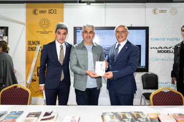 First image displays a group of formally dressed men and women including some in headscarves standing around a table covered with stacks of blue-covered books at an indoor event space with Turkish flags and banners overhead. Second image shows three men in suits one holding a book posing in front of a banner for a book model event with a screen and books on a table. Third image features a group of young men and women some in headscarves standing together at a booth labeled for second category fair with coffee cups and displayed items on the table. Fourth image depicts a line of children in colorful traditional Turkish attire including embroidered dresses and vests flanked by adult men in suits and traditional headwear in an indoor hall with modern lighting.
