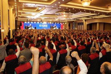 First image shows a large indoor conference hall with a stage featuring Turkish flags and a screen displaying a speaker, surrounded by a massive crowd of attendees mostly wearing red and black vests, many raising their fists in unity. Second image depicts a man in a red and black vest standing at a white podium with a microphone, pointing upwards with his index finger, against a blue backdrop with text reading Buyuk Bulusma 1 Kasim 2025 and star motifs. Third image captures rows of formally dressed men and women standing shoulder to shoulder in a grand hall with blue banners reading Buyuk Bulusma, applauding or raising hands. Fourth image illustrates a wide view of a spacious venue with multiple rows of seated and standing participants in formal attire, facing a stage with blue banners inscribed with Buyuk Bulusma text, floral decorations on the floor.