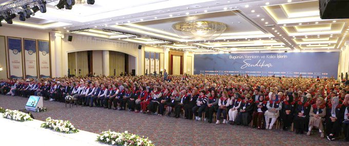 First image shows a large indoor conference hall with a stage featuring Turkish flags and a screen displaying a speaker, surrounded by a massive crowd of attendees mostly wearing red and black vests, many raising their fists in unity. Second image depicts a man in a red and black vest standing at a white podium with a microphone, pointing upwards with his index finger, against a blue backdrop with text reading Buyuk Bulusma 1 Kasim 2025 and star motifs. Third image captures rows of formally dressed men and women standing shoulder to shoulder in a grand hall with blue banners reading Buyuk Bulusma, applauding or raising hands. Fourth image illustrates a wide view of a spacious venue with multiple rows of seated and standing participants in formal attire, facing a stage with blue banners inscribed with Buyuk Bulusma text, floral decorations on the floor.