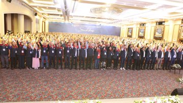 First image shows a large indoor conference hall with a stage featuring Turkish flags and a screen displaying a speaker, surrounded by a massive crowd of attendees mostly wearing red and black vests, many raising their fists in unity. Second image depicts a man in a red and black vest standing at a white podium with a microphone, pointing upwards with his index finger, against a blue backdrop with text reading Buyuk Bulusma 1 Kasim 2025 and star motifs. Third image captures rows of formally dressed men and women standing shoulder to shoulder in a grand hall with blue banners reading Buyuk Bulusma, applauding or raising hands. Fourth image illustrates a wide view of a spacious venue with multiple rows of seated and standing participants in formal attire, facing a stage with blue banners inscribed with Buyuk Bulusma text, floral decorations on the floor.