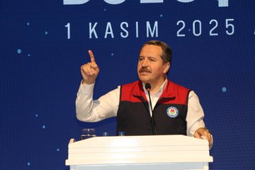 First image shows a large indoor conference hall with a stage featuring Turkish flags and a screen displaying a speaker, surrounded by a massive crowd of attendees mostly wearing red and black vests, many raising their fists in unity. Second image depicts a man in a red and black vest standing at a white podium with a microphone, pointing upwards with his index finger, against a blue backdrop with text reading Buyuk Bulusma 1 Kasim 2025 and star motifs. Third image captures rows of formally dressed men and women standing shoulder to shoulder in a grand hall with blue banners reading Buyuk Bulusma, applauding or raising hands. Fourth image illustrates a wide view of a spacious venue with multiple rows of seated and standing participants in formal attire, facing a stage with blue banners inscribed with Buyuk Bulusma text, floral decorations on the floor.