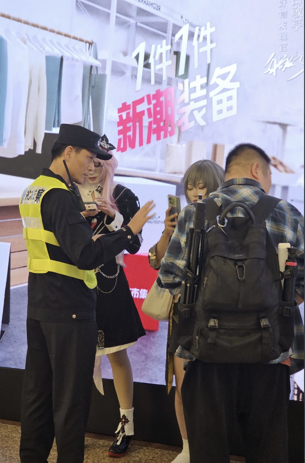一群人站在地铁站区，背景是大广告屏，用汉字显示宣传文字。两名身穿黄色背心和深色制服的警察在场，一名将手机放在耳边，同时用手比划，另一名站在附近。两个年轻人，一名穿着格子衬衫和背包的短发男性，以及一名穿着黑色连衣裙和白色袜子的长发女性，似乎是互动的焦点，女性拿着手机。从后面可以看到另一个背着背包的人。场景包括衣架和带有中文文字的红色横幅。