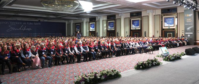 First image shows a man in a red and white vest standing at a blue podium pointing emphatically during a speech, with a large blue banner behind reading 20 Turkıyeye Buluşması 1 Kasım 2025 Antalya and Memur-Sen logo. Second image depicts a large audience of people in formal attire seated in rows on a carpeted floor in a grand hall with blue banners reading Sendika and floral decorations. Third image captures another crowded conference room with attendees in suits and dresses seated under chandeliers and blue banners reading Sendika. Fourth image features the same man from behind at a podium addressing a vast seated audience in a spacious hall with blue Sendika banners and floral arrangements.