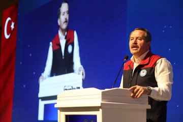 First image shows a man in a white shirt and red-and-white vest speaking at a white podium with a microphone, standing in front of a large screen displaying a similar image of himself, flanked by Turkish flags on blue and red backgrounds in a conference setting. Second image depicts a large seated audience of men and women in formal attire, many wearing red sashes, in a grand hall with blue walls, chandeliers, and banners reading Sendika and a logo, arranged in rows facing a stage with floral decorations. Third image features a white book cover titled EMEK MÜCADELEMİZDE 1124 KAZANIMIZ with images of cheering crowds, a QR code, and logos of Memur-Sen and Eğitim Bir-Sen, presented at an angle on a surface. Fourth image shows a diverse group of men and women in suits and vests, some with red sashes, seated and clapping enthusiastically in a hall with banners reading İSTANBUL SİVAS ANKARA on pillars and a small table with flowers nearby.