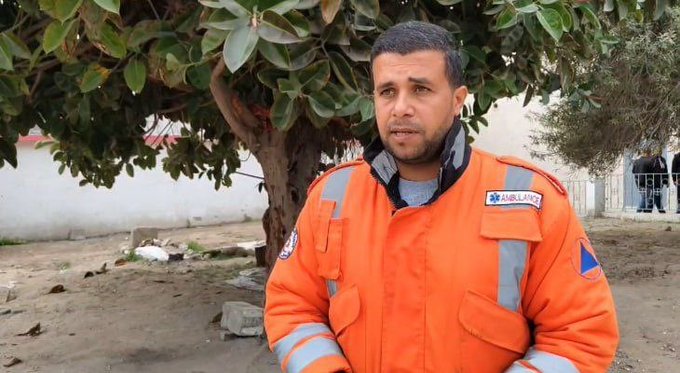 A man with short dark hair and a beard stands outdoors wearing an orange high-visibility jacket with reflective strips and a badge labeled Civil Defence over a blue shirt. He has a serious expression and is positioned in front of a large green tree with broad leaves and a white wall in the background. Debris is scattered on the ground nearby and other people are visible in the distance wearing similar uniforms.