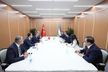 First image shows two officials, one male in a dark suit holding a red folder with Turkish emblem and one female in a blue suit holding a blue folder, shaking hands in front of Turkish and Uzbek flags against a wooden backdrop. Second image depicts a group of suited officials seated around a long white table in a modern room with wooden walls, Turkish and Uzbek flags, water bottles, and plants, engaged in discussion. Third image captures officials at a similar table, with one male official signing a document on red paper and a female official writing on blue paper, surrounded by flags, water bottles, and chairs.