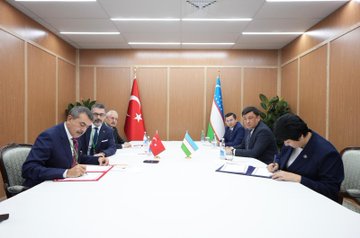 First image shows two officials, one male in a dark suit holding a red folder with Turkish emblem and one female in a blue suit holding a blue folder, shaking hands in front of Turkish and Uzbek flags against a wooden backdrop. Second image depicts a group of suited officials seated around a long white table in a modern room with wooden walls, Turkish and Uzbek flags, water bottles, and plants, engaged in discussion. Third image captures officials at a similar table, with one male official signing a document on red paper and a female official writing on blue paper, surrounded by flags, water bottles, and chairs.