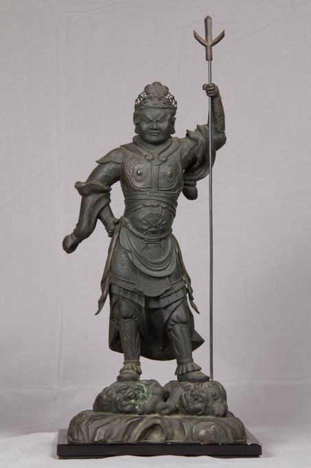 Bronze statue of Bishamonten standing on a base with demon heads holding a trident in the left hand with the right hand on the hip wearing armor and a crown with a fierce expression stepping on evil spirits