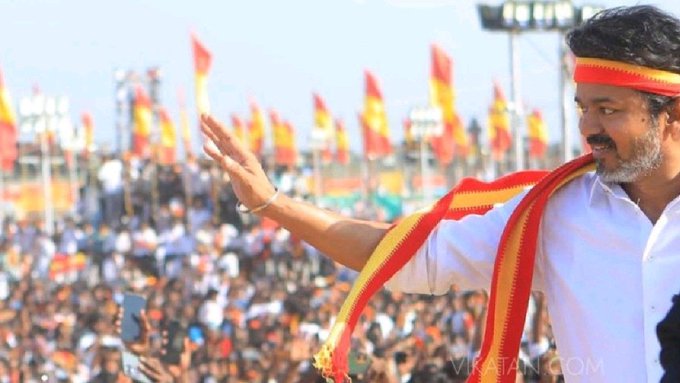 A man with a beard wearing a white shirt and red and yellow scarf stands on a stage raising his right hand in a gesture with orange flags and a crowd in the background at an outdoor event under stadium lights