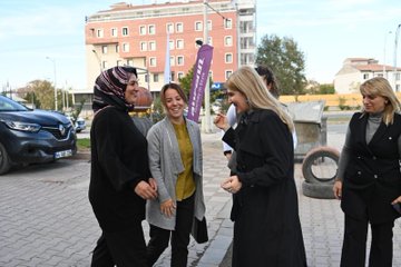 First image shows five women standing in a tire shop surrounded by stacked tires and shelves with automotive products one woman in a headscarf holds a long rectangular plaque while others wear professional attire like vests and coats with a large banner reading Süt in the background. Second image depicts six women outdoors near a building under construction chatting and smiling some wearing headscarves and coats with a purple banner and parked cars visible nearby. Third image features six women seated around a wooden desk in an office setting with automotive posters on walls one in headscarf others in coats and vests discussing with items like a phone and bottle on the table.