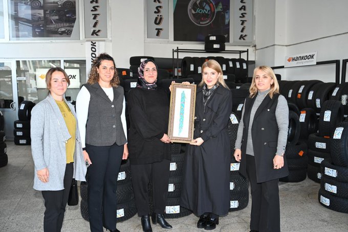 First image shows five women standing in a tire shop surrounded by stacked tires and shelves with automotive products one woman in a headscarf holds a long rectangular plaque while others wear professional attire like vests and coats with a large banner reading Süt in the background. Second image depicts six women outdoors near a building under construction chatting and smiling some wearing headscarves and coats with a purple banner and parked cars visible nearby. Third image features six women seated around a wooden desk in an office setting with automotive posters on walls one in headscarf others in coats and vests discussing with items like a phone and bottle on the table.