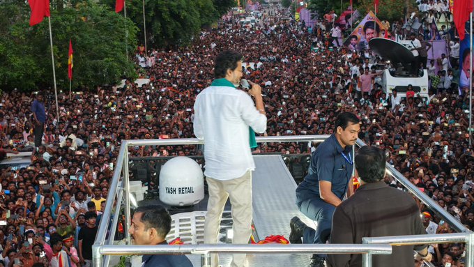 A man in a white shirt and green shawl stands on a metal platform speaking into a microphone with another man in a blue shirt nearby. A large crowd of people gathers below in an open area surrounded by trees red flags and vehicles. A white satellite dish is mounted on the platform and banners are visible in the background.
