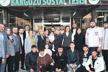First image shows a large group of adults and children posing in front of a building entrance with a green sign reading Barguzu Sosyal Tesis, some holding a baby, all dressed in casual and formal winter clothing. Second image depicts four women seated at a wooden table with plates of food, glasses of water, and desserts, one child in a pink hat sitting among them, background showing windows and blurred people. Third image features a group of men and women around a table with food items like pastries and drinks, a blue banner with Turkish text for youth center, some wearing headscarves, gesturing during discussion. Fourth image captures another group of adults and a woman holding a baby outside a building entrance with plants and lights, all in coats and jackets.
