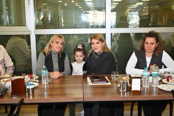 First image shows a large group of adults and children posing in front of a building entrance with a green sign reading Barguzu Sosyal Tesis, some holding a baby, all dressed in casual and formal winter clothing. Second image depicts four women seated at a wooden table with plates of food, glasses of water, and desserts, one child in a pink hat sitting among them, background showing windows and blurred people. Third image features a group of men and women around a table with food items like pastries and drinks, a blue banner with Turkish text for youth center, some wearing headscarves, gesturing during discussion. Fourth image captures another group of adults and a woman holding a baby outside a building entrance with plants and lights, all in coats and jackets.