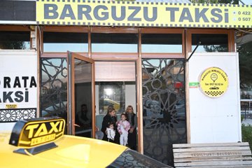 First image shows a group of about ten adults including several women in professional attire and men in casual jackets standing together in front of a yellow taxi stand vehicle labeled Barguzu Taksi with traditional Turkish conical roofs and satellite dish a young girl in pink dress stands among them smiling with trees in the background. Second image depicts five people seated on a light blue couch in an indoor setting with beige walls and a mirror one man in black tracksuit another in jeans and black top three women in dark clothing and a young girl in pink outfit with bow headband holding toys looking at the camera. Third image features the exterior of the Barguzu Taksi building with yellow signage phone number and taxi symbol open wooden door showing interior a yellow taxi parked nearby with several people including women and children standing outside wooden bench and patterned wall decorations visible.