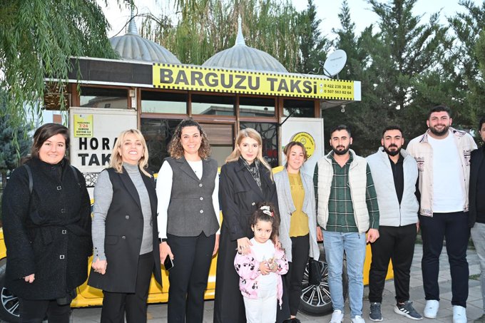 First image shows a group of about ten adults including several women in professional attire and men in casual jackets standing together in front of a yellow taxi stand vehicle labeled Barguzu Taksi with traditional Turkish conical roofs and satellite dish a young girl in pink dress stands among them smiling with trees in the background. Second image depicts five people seated on a light blue couch in an indoor setting with beige walls and a mirror one man in black tracksuit another in jeans and black top three women in dark clothing and a young girl in pink outfit with bow headband holding toys looking at the camera. Third image features the exterior of the Barguzu Taksi building with yellow signage phone number and taxi symbol open wooden door showing interior a yellow taxi parked nearby with several people including women and children standing outside wooden bench and patterned wall decorations visible.