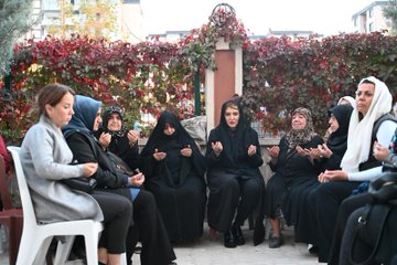 First image shows a group of women mostly wearing hijabs and abayas sitting in a circle on chairs outdoors against a red vine-covered wall with one woman holding hands prayerfully. Second image depicts several women and two men in casual attire embracing and standing together near a red building wall with one woman in a black lace headscarf hugging another. Third image features a man holding a microphone speaking to a seated group of women in hijabs and abayas around white chairs in an outdoor paved area with red vines in the background. Fourth image portrays women in hijabs including one with a blue headscarf and men in jackets standing and talking near a red wall with buildings visible.