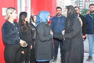 First image shows a group of women mostly wearing hijabs and abayas sitting in a circle on chairs outdoors against a red vine-covered wall with one woman holding hands prayerfully. Second image depicts several women and two men in casual attire embracing and standing together near a red building wall with one woman in a black lace headscarf hugging another. Third image features a man holding a microphone speaking to a seated group of women in hijabs and abayas around white chairs in an outdoor paved area with red vines in the background. Fourth image portrays women in hijabs including one with a blue headscarf and men in jackets standing and talking near a red wall with buildings visible.