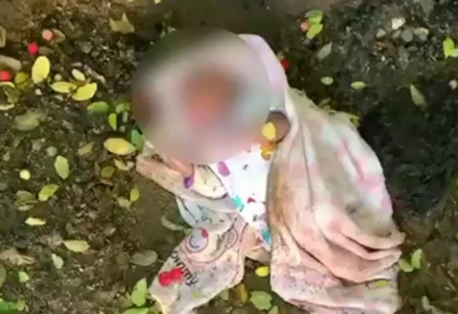 A blurred image of an infant wrapped in light-colored clothing with colorful patterns lying on the ground covered in dirt and scattered yellow and red fallen leaves in an outdoor setting.