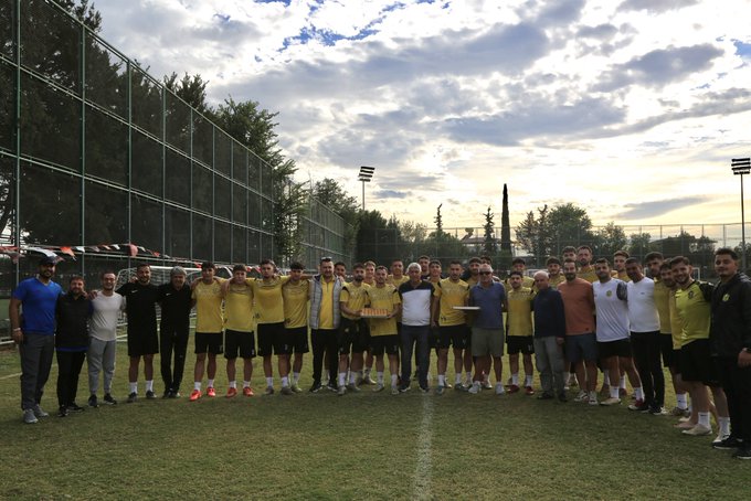 Group of male athletes in yellow jerseys and shorts stand on a grassy soccer field surrounded by a chain-link fence, with several men in casual clothes including white shirts and pants joining them for a team photo. Floodlights and trees are visible in the background under a partly cloudy sky at dusk. Another similar group photo shows players and supporters posing together on the field with similar attire and setting elements.